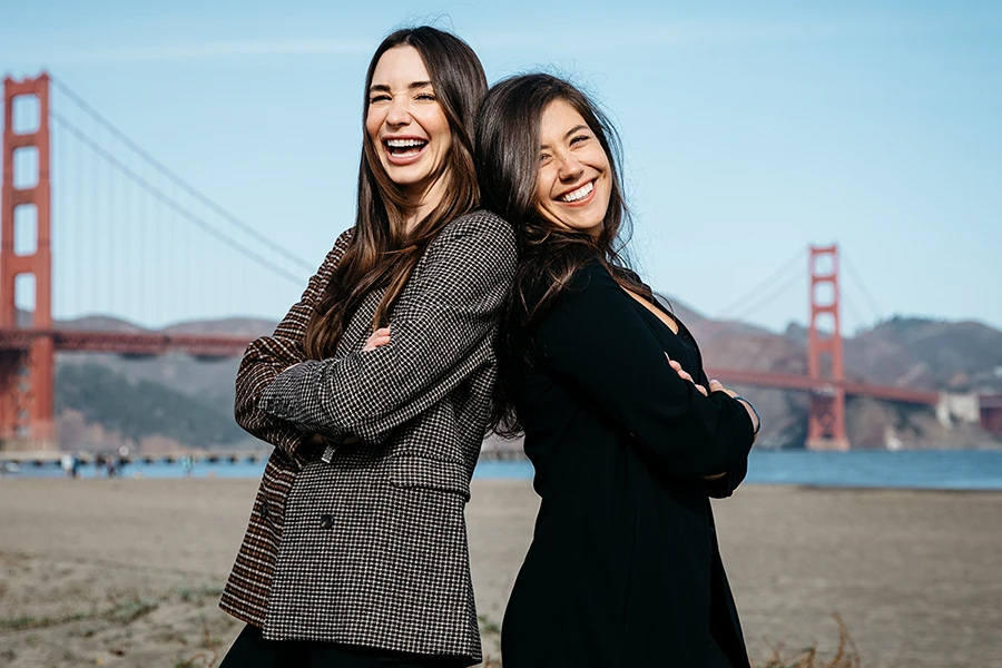 Tara in a navy blazer, standing back to back with Blaine, outside on the beach in San Francisco, with the Golden Gate Bridge in the background