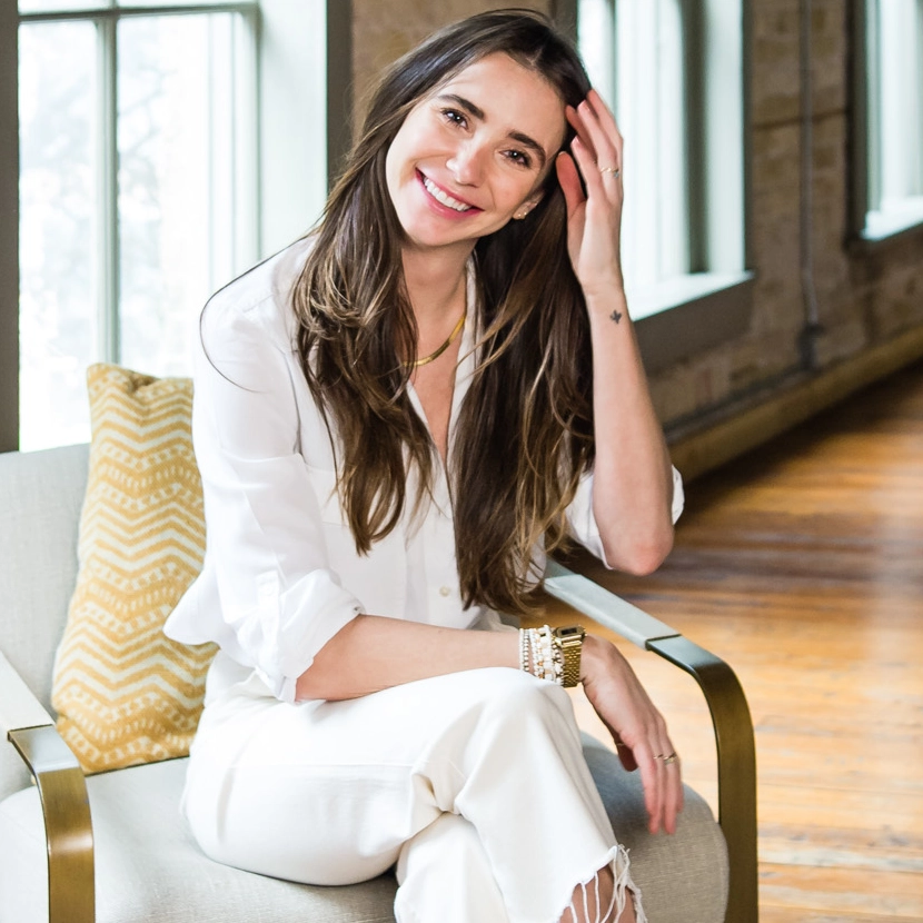 Photo of Blaine in a white outfit in a loft-like building, smiling, and touching her hair