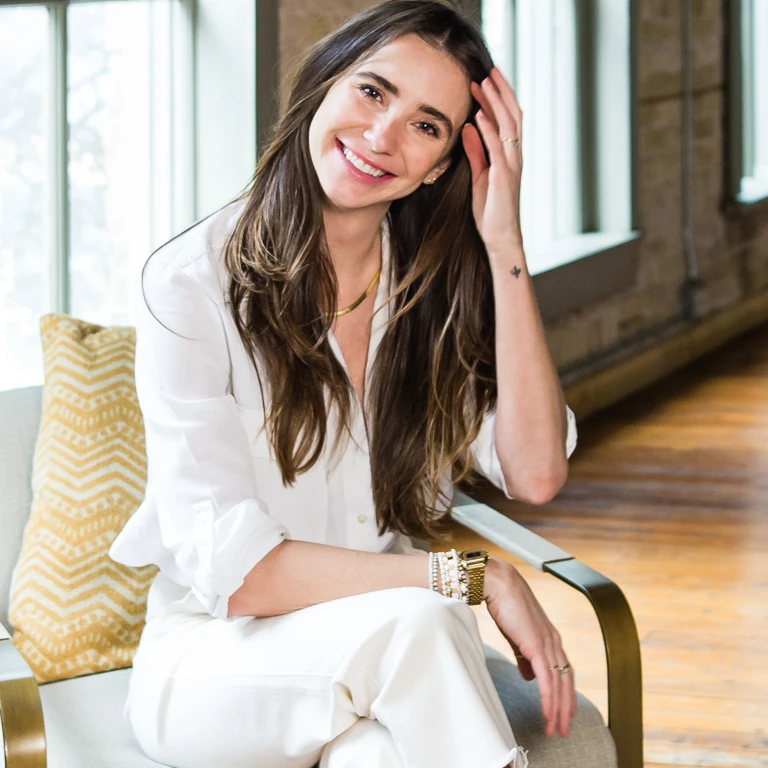 Photo of Blaine in a white outfit in a loft-like building, seated, smiling, touching her hair.